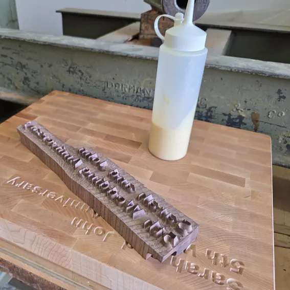 Walnut inlay lettering reading "Sarah and John, 5th Wedding Anniversary" being fitted into a personalised end-grain maple chopping board